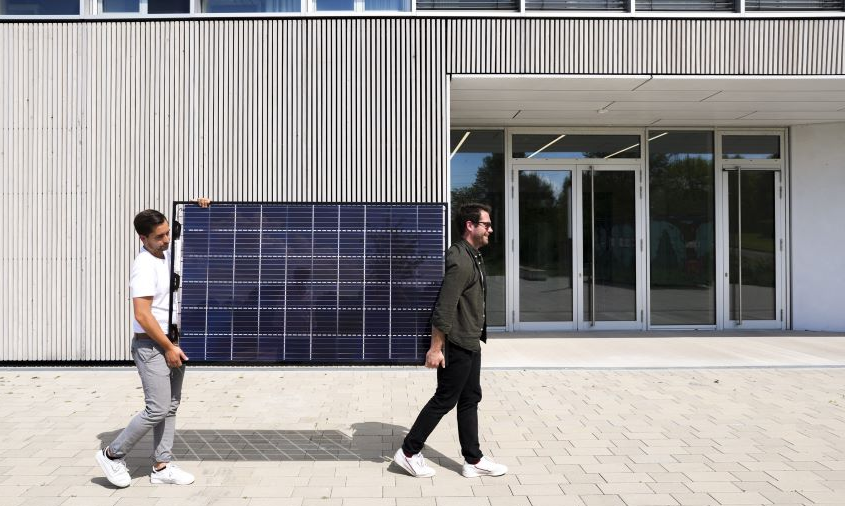 Zwei junge Männer tragen ein Photovoltaik-Modul über einen Platz vor einem Gebäude 